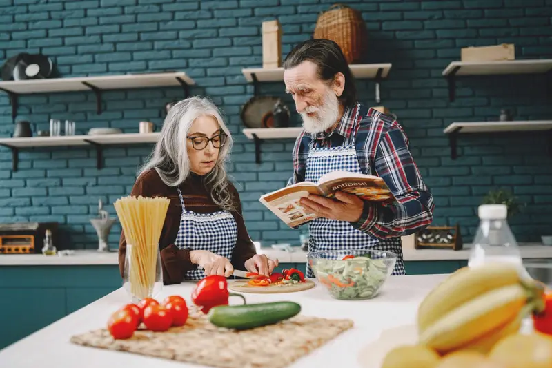 An older couple cooks a fresh meal together, reading a recipe book